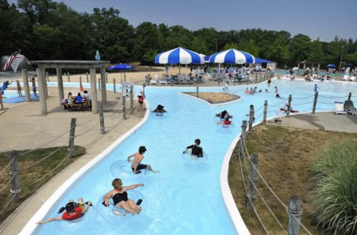 A sunny water park scene with people floating on tubes in a lazy river, surrounded by trees and blue umbrellas.