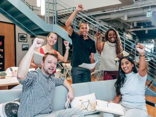 A diverse group of five people in an office, smiling and raising their fists in celebration, holding papers.