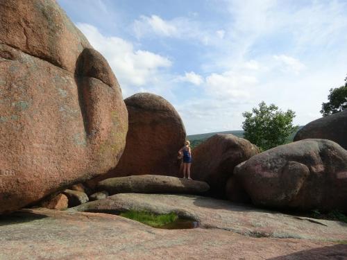 A person stands on large, weathered rocks under a blue sky with scattered clouds, surrounded by greenery.