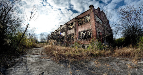 Abandoned brick building surrounded by overgrown vegetation and a cloudy sky.
