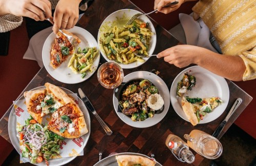 A table filled with various dishes, including pizza, pasta, and salad, with hands reaching for the food.