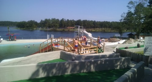 A water park scene with a slide, mini-golf area, and people enjoying the sunny day by a lake surrounded by trees.
