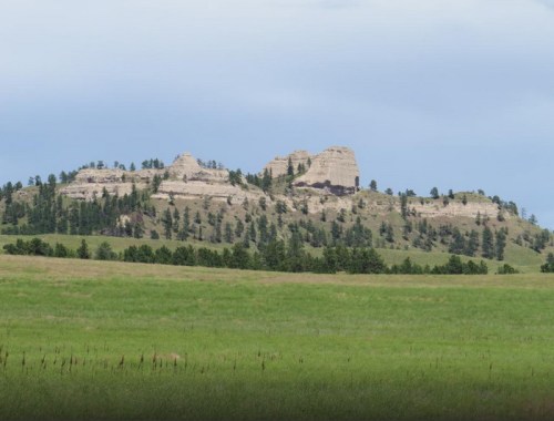 A rocky hilltop rises above a green field, surrounded by trees under a cloudy sky.