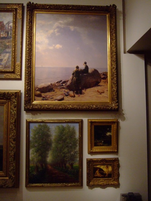 A gallery wall featuring several framed paintings, including landscapes and a couple by the sea.