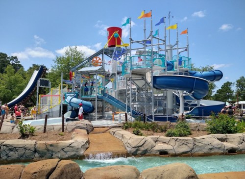 A colorful water park with slides, pools, and flags, surrounded by trees and visitors enjoying the sunny day.