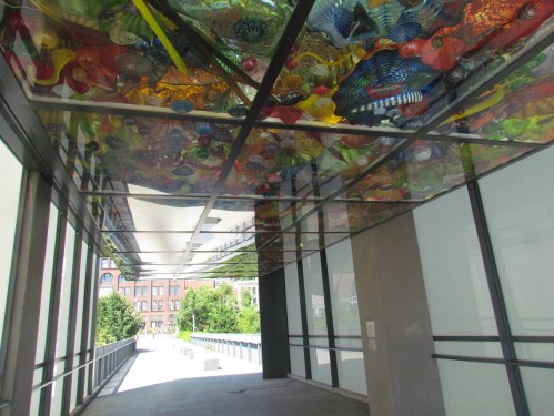 A glass ceiling adorned with colorful glass art, viewed from a modern walkway with natural light streaming in.