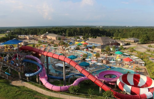 Aerial view of a vibrant water park featuring slides, pools, and colorful umbrellas, surrounded by greenery.