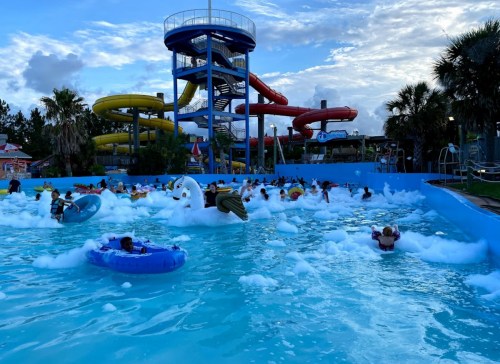 A crowded water park wave pool with colorful slides and foam, featuring people on floats enjoying the water.