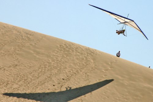 A hang glider soars above a sandy dune, casting a large shadow on the ground below.