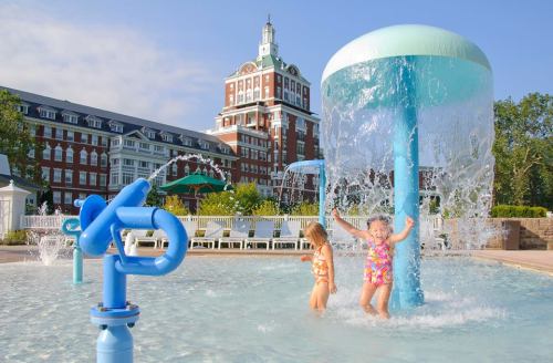 Two children play in a splash pad with water features, set against a large hotel in the background on a sunny day.
