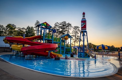 Colorful water slides and a lighthouse structure at a water park, with children playing in the pool at sunset.