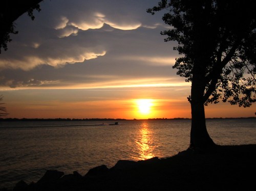 Sunset over a calm lake, with silhouetted trees and clouds reflecting warm colors in the sky.