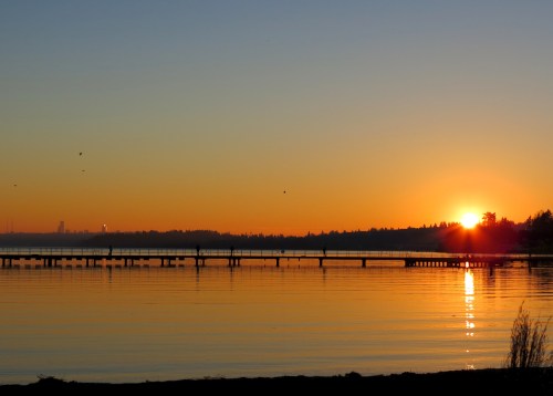 A serene sunset over a calm lake, with a silhouette of a pier and trees against an orange and blue sky.