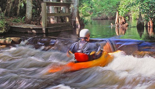 A person in an orange kayak navigates a rapid river surrounded by lush greenery and trees.
