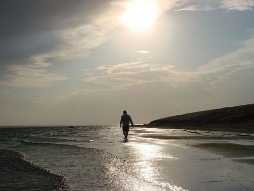 A person walks along a beach at sunset, with shimmering water and a cloudy sky in the background.
