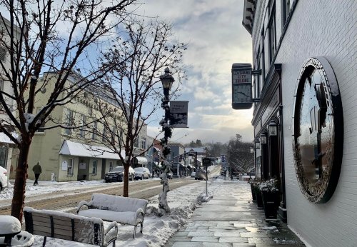 A snowy street scene with trees, benches, and buildings under a cloudy sky, creating a serene winter atmosphere.