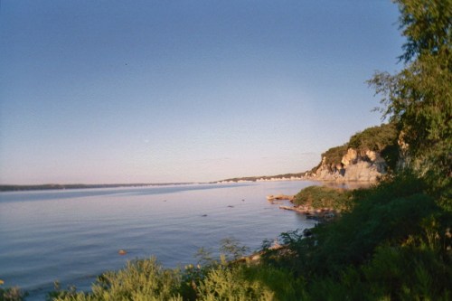 A serene river scene with calm waters, lush greenery, and rocky cliffs under a clear blue sky.