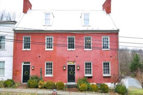 A red brick house with a white roof, featuring multiple windows and a front garden, set along a street.