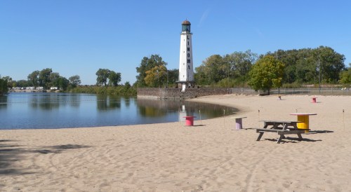 A sandy beach with colorful picnic tables, a calm lake, and a tall white lighthouse in the background under a clear blue sky.