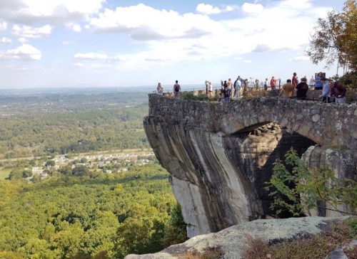 A scenic overlook with people standing on a stone ledge, overlooking a lush valley and distant hills under a blue sky.