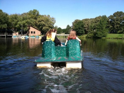 Two people paddle a green pedal boat on a calm lake, surrounded by trees and a building in the background.