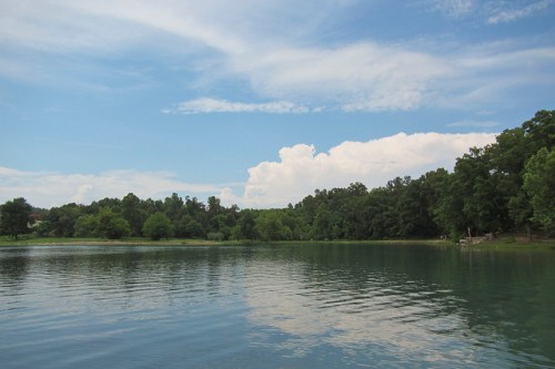 A serene lake surrounded by lush greenery and a clear blue sky with fluffy clouds.