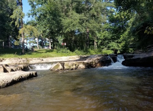 A serene river scene with gentle flowing water, rocky banks, and lush green trees in the background.