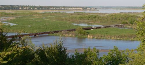 A scenic view of a river with a wooden bridge, surrounded by lush greenery and distant hills under a clear sky.