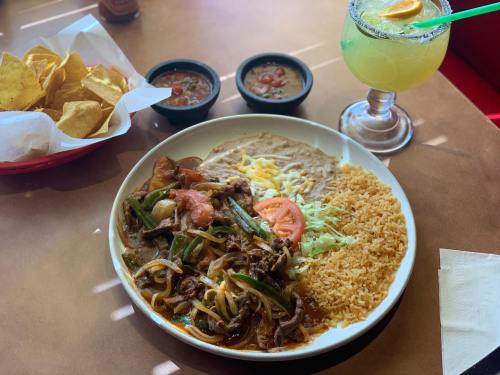 A plate of sizzling beef fajitas with rice, refried beans, and fresh toppings, accompanied by chips, salsa, and a margarita.