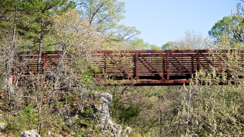 A wooden bridge spans a rocky area, surrounded by lush greenery and trees under a clear blue sky.
