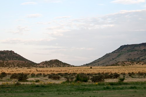 A wide landscape featuring two low hills, grassy plains, and a cloudy sky in the background.