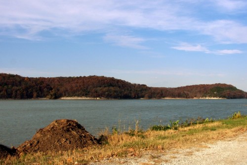 A serene lake surrounded by hills with autumn foliage under a clear blue sky. A gravel path runs along the shore.