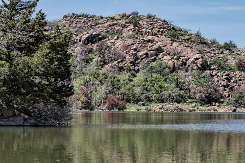 A serene lake surrounded by rocky hills and lush greenery under a clear blue sky. Reflections shimmer on the water.