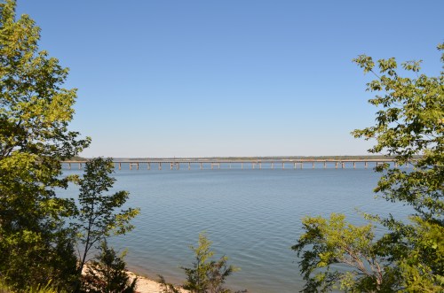A calm lake with a clear blue sky, framed by trees, and a long bridge stretching across the water in the distance.