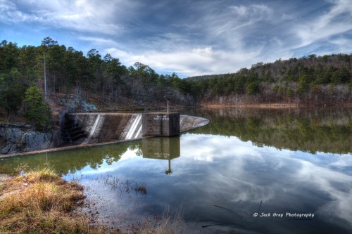 A serene landscape featuring a dam reflected in calm water, surrounded by trees and a cloudy sky.