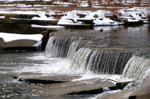 A serene waterfall cascades over rocky ledges, surrounded by snow and calm water.