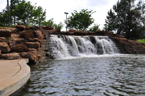 A serene waterfall cascades over rocks into a calm pond, surrounded by greenery and trees.
