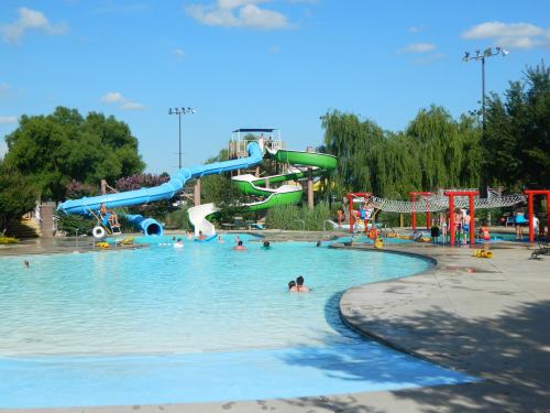 A sunny water park scene featuring a large pool, water slides, and people enjoying the water.