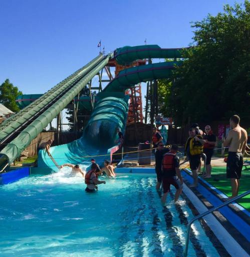 A water park scene with a large slide, people splashing in the pool, and others waiting nearby in swimwear.