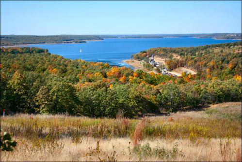 A scenic view of a lake surrounded by colorful autumn trees and rolling hills under a clear blue sky.
