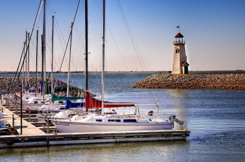 A marina with sailboats docked, featuring a lighthouse on a rocky pier under a clear blue sky.