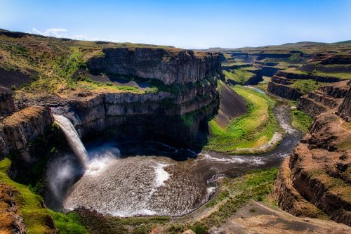 A stunning waterfall cascades into a river, surrounded by steep cliffs and lush green landscapes under a clear blue sky.