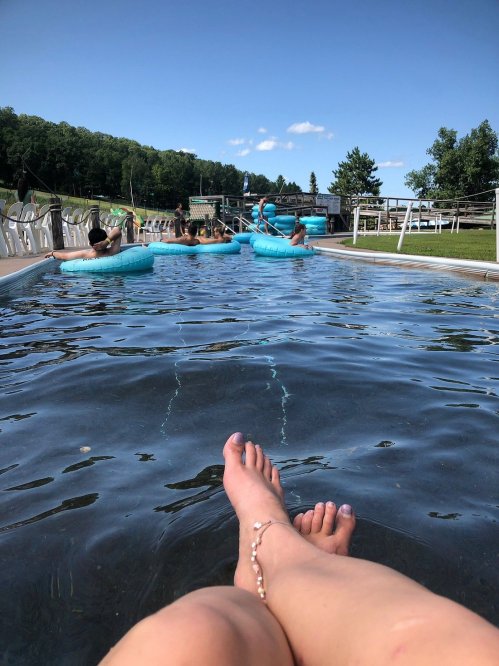 A view of a pool with people floating on inner tubes, and a person's feet resting at the edge of the water.