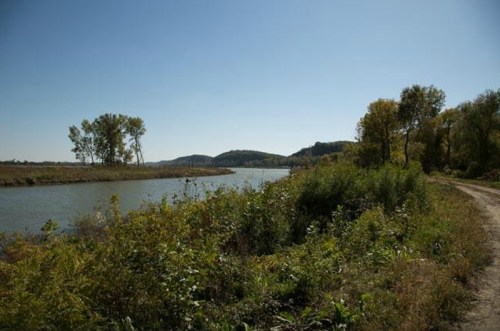 A serene river scene with trees lining the banks and rolling hills in the background under a clear blue sky.