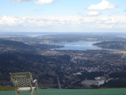 A scenic view from a mountain overlooking a lake and town, with a white chair in the foreground against a cloudy sky.