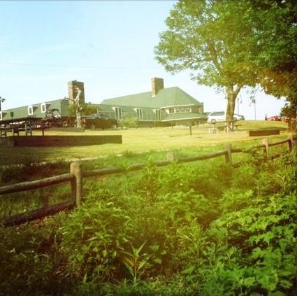 A green-roofed building surrounded by grass and trees, with a wooden fence in the foreground.