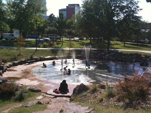 Children play in a splash pad surrounded by greenery, with a few adults relaxing nearby on a sunny day.