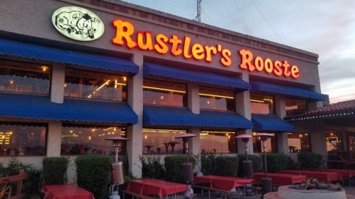 Exterior of Rustler's Rooste restaurant with bright signage and outdoor seating under blue awnings at sunset.