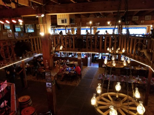 A rustic restaurant interior with wooden beams, hanging lights, and diners on multiple levels.