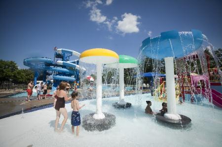 Children play in a splash pad with colorful water features, while water slides and other attractions are in the background.
